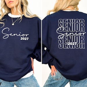 May include: Navy blue sweatshirt with the word "Senior" in white script and the year "2027" printed on the front. The back of the sweatshirt repeats "Senior 2027" in a stacked design. The sweatshirt has a white crew neck.