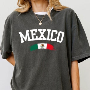May include: A dark gray t-shirt with a white graphic of the word "MEXICO" in an arch with a stylized Mexican flag underneath.