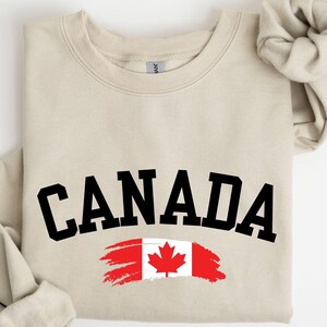 May include: A beige sweatshirt with the word "CANADA" in black letters across the front. The word is in an arc shape with a Canadian flag painted in red and white with a red maple leaf in the center.