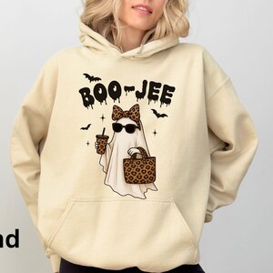 Op de afbeelding: Zandkleurige hoodie met een afbeelding van een spook met een zonnebril en een luipaardprint strik, met een koffiekopje en een luipaardprint tas. De tekst "BOO-JEE" staat boven het spook, met vleermuizen en sterren.