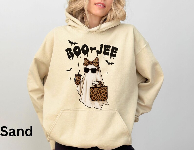 Op de afbeelding: Zandkleurige hoodie met een afbeelding van een spook met een zonnebril en een luipaardprint strik, met een koffiekopje en een luipaardprint tas. De tekst "BOO-JEE" staat boven het spook, met vleermuizen en sterren.