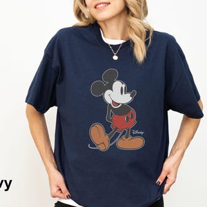 May include: Navy blue t-shirt featuring a classic Mickey Mouse graphic. The cartoon character is depicted in his iconic pose, wearing red shorts and brown shoes. The word "Disney" is printed below the character. The word "Navy" is printed in the bottom left corner.