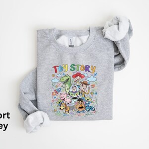 May include: Sport grey sweatshirt featuring a colorful graphic of Toy Story characters, including Woody, Buzz Lightyear, and Rex. The words "Toy Story" are written above the characters in a playful font. The sweatshirt has a crew neck and long sleeves.