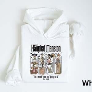 May include: White hooded sweatshirt featuring a graphic from The Haunted Mansion. The graphic includes the text "The Haunted Mansion" and "Welcome Foolish Mortals Est. 1969" with cartoon characters. The sweatshirt has long sleeves and a drawstring hood.