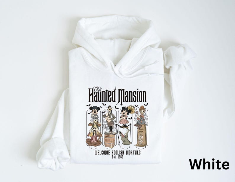 May include: White hooded sweatshirt featuring a graphic from The Haunted Mansion. The graphic includes the text "The Haunted Mansion" and "Welcome Foolish Mortals Est. 1969" with cartoon characters. The sweatshirt has long sleeves and a drawstring hood.