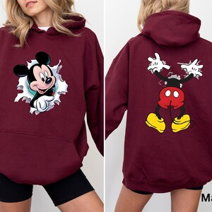 May include: Maroon hoodie featuring a Mickey Mouse graphic on the front and back. The front design shows Mickey breaking through a torn paper effect. The back displays Mickey's arms and legs in a similar style. The hoodie has a kangaroo pocket and a hood.