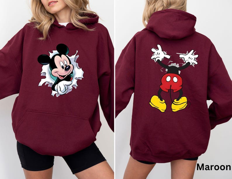 May include: Maroon hoodie featuring a Mickey Mouse graphic on the front and back. The front design shows Mickey breaking through a torn paper effect. The back displays Mickey's arms and legs in a similar style. The hoodie has a kangaroo pocket and a hood.