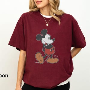 May include: Maroon t-shirt featuring a classic Mickey Mouse graphic. The design shows Mickey in his iconic pose, with red shorts, brown shoes, and black gloves. The shirt has a white collar and the Disney logo.
