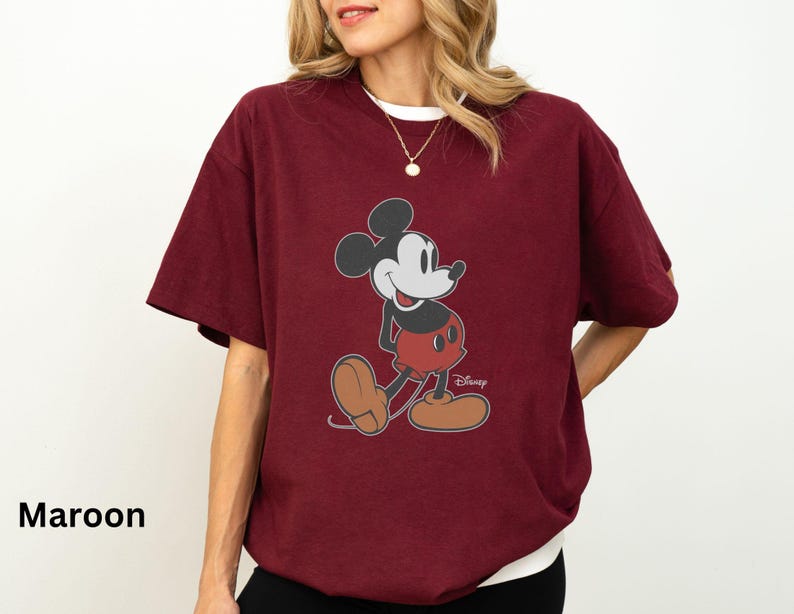 May include: Maroon t-shirt featuring a classic Mickey Mouse graphic. The design shows Mickey in his iconic pose, with red shorts, brown shoes, and black gloves. The shirt has a white collar and the Disney logo.