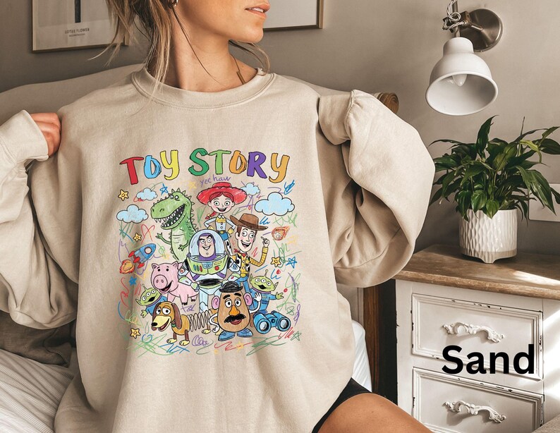 May include: A sand-colored sweatshirt featuring a colorful graphic of characters from the animated film "Toy Story." The design includes the title "Toy Story" in a rainbow font, along with images of Woody, Buzz Lightyear, and other characters.