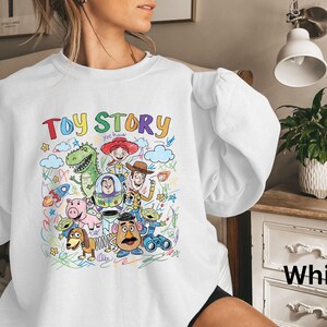 May include: White sweatshirt featuring a colorful graphic of characters from the animated film "Toy Story." The design includes the words "Toy Story" in rainbow colors, along with images of Woody, Buzz Lightyear, and other characters.