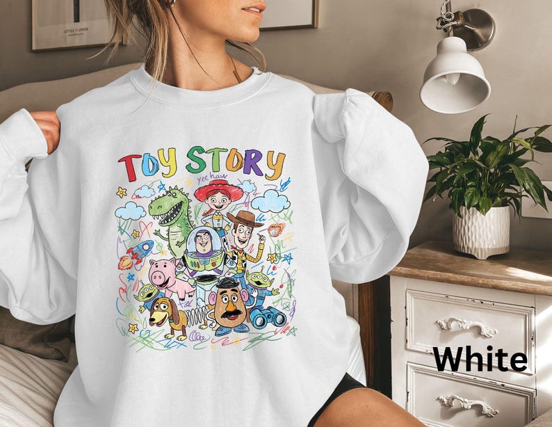 May include: White sweatshirt featuring a colorful graphic of characters from the animated film "Toy Story." The design includes the words "Toy Story" in rainbow colors, along with images of Woody, Buzz Lightyear, and other characters.