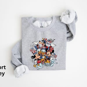 Puede incluir: Sudadera gris deportiva con un dise&ntilde;o de Mickey Mouse, Minnie Mouse, Donald, Goofy y Pluto con atuendos patri&oacute;ticos. El dise&ntilde;o incluye banderas americanas y el texto "AMERICA 250th ANNIVERSARY". La sudadera tiene cuello redondo y mangas largas.