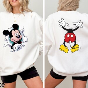 May include: White sweatshirt featuring a Mickey Mouse graphic on the front and back. The front design shows Mickey breaking through a torn-paper effect. The back design depicts Mickey's legs and arms. The word "White" is printed at the bottom.