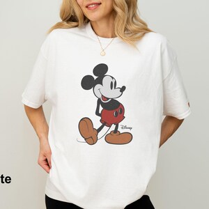 May include: White t-shirt featuring a classic Mickey Mouse graphic. The cartoon character is depicted in black, red, and brown, with the Disney logo visible. The word "White" is printed at the bottom left of the image.