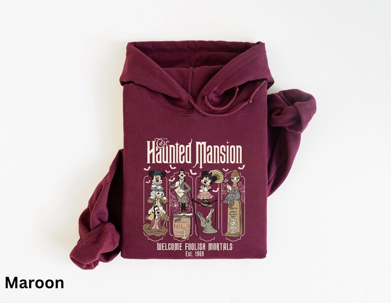 May include: Maroon hoodie featuring a graphic from the Haunted Mansion ride. The design includes characters and the text "The Haunted Mansion" and "Welcome Foolish Mortals Est. 1969". The hoodie has a hood and long sleeves.
