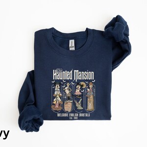 May include: Navy blue sweatshirt featuring a graphic from the Haunted Mansion ride. The design includes the text "The Haunted Mansion" and "Welcome Foolish Mortals Est. 1969" with cartoon characters. The sweatshirt is made of a soft material.