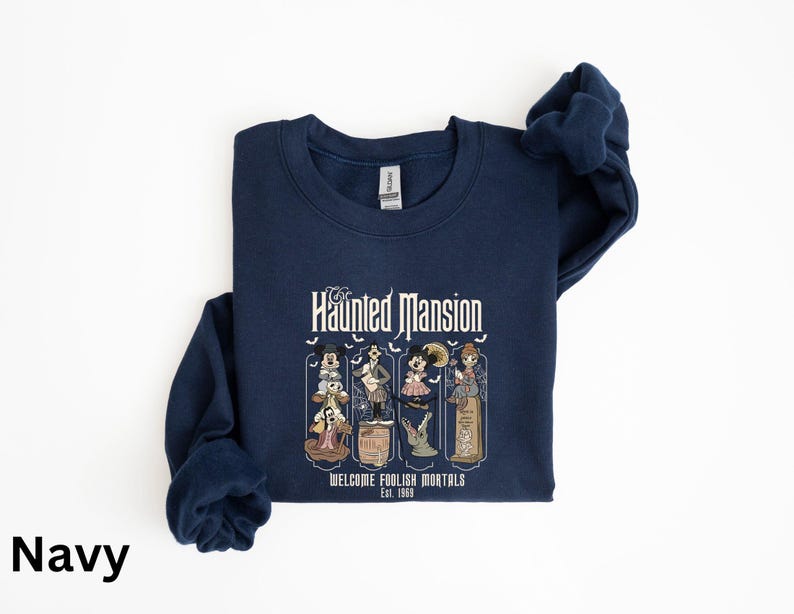 May include: Navy blue sweatshirt featuring a graphic from the Haunted Mansion ride. The design includes the text "The Haunted Mansion" and "Welcome Foolish Mortals Est. 1969" with cartoon characters. The sweatshirt is made of a soft material.