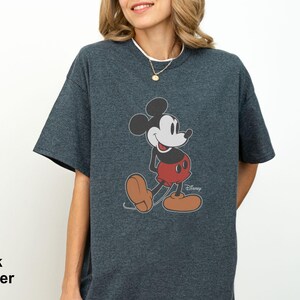 May include: Dark heather gray t-shirt featuring a classic Mickey Mouse graphic. The cartoon character is depicted in his iconic pose, wearing red shorts, brown shoes, and white gloves. The Disney logo is visible. The words "Dark Heather" are printed on the bottom left.