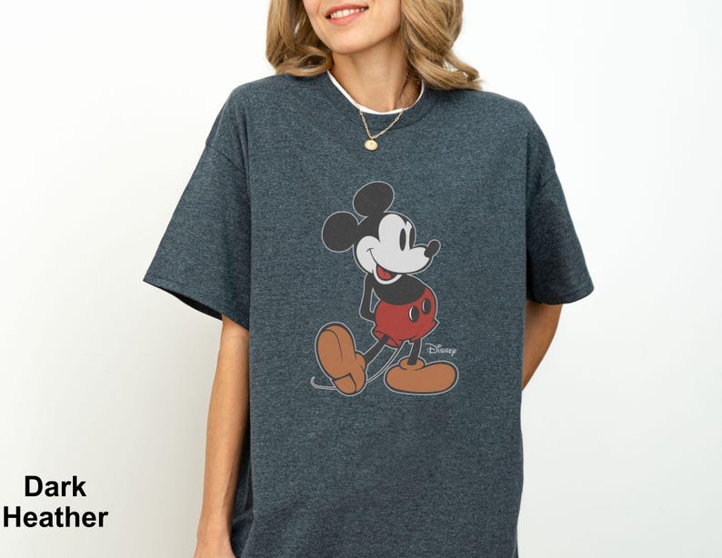 May include: Dark heather gray t-shirt featuring a classic Mickey Mouse graphic. The cartoon character is depicted in his iconic pose, wearing red shorts, brown shoes, and white gloves. The Disney logo is visible. The words "Dark Heather" are printed on the bottom left.