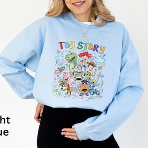 May include: Light blue sweatshirt featuring a colorful graphic of characters from the animated film "Toy Story." The design includes Woody, Buzz Lightyear, Rex, and other characters. The words "Toy Story" are written in a playful font above the characters.