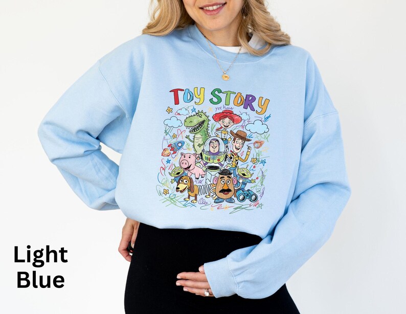 May include: Light blue sweatshirt featuring a colorful graphic of characters from the animated film "Toy Story." The design includes Woody, Buzz Lightyear, Rex, and other characters. The words "Toy Story" are written in a playful font above the characters.