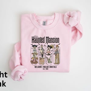 May include: Light pink sweatshirt featuring a graphic from the Haunted Mansion ride. The design includes cartoon characters and the text "Haunted Mansion" and "Welcome Foolish Mortals Est. 1969".