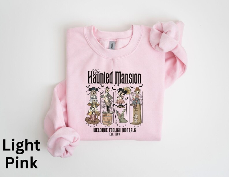 May include: Light pink sweatshirt featuring a graphic from the Haunted Mansion ride. The design includes cartoon characters and the text "Haunted Mansion" and "Welcome Foolish Mortals Est. 1969".