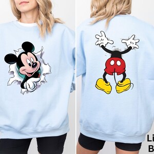 May include: Light blue sweatshirt featuring Mickey Mouse graphics on the front and back. The front graphic shows Mickey breaking through a torn surface, while the back displays Mickey's full body. The text "Light Blue" is visible.