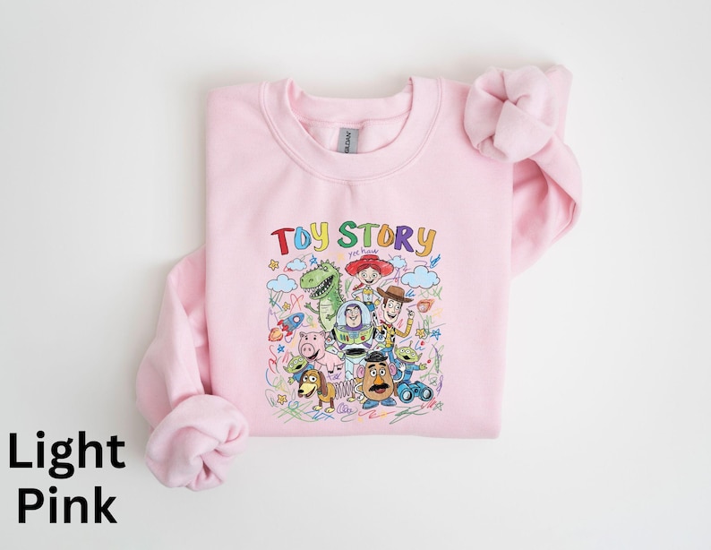 May include: Light pink sweatshirt featuring a colorful graphic of characters from the animated film "Toy Story." The text "Toy Story" is displayed above the characters. The sweatshirt has long sleeves and a crew neck.