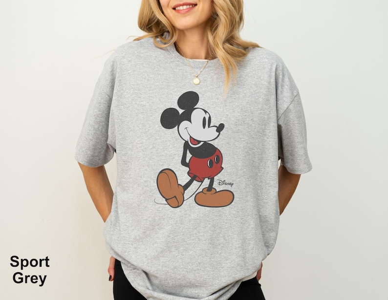 May include: Sport grey t-shirt featuring a classic Mickey Mouse graphic. The cartoon character is depicted in his iconic pose, wearing red shorts, brown shoes, and white gloves. The Disney logo is visible. The shirt is a light grey color.