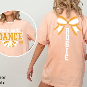 May include: Heather peach t-shirt with a large bow graphic on the back and the name "Rosie" printed vertically. The front features the text "Team Name Dance 2025" in gold and white.
