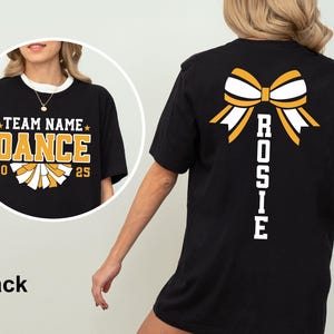 May include: Black t-shirt with a white collar featuring a gold and white bow design on the back. The front of the shirt has the text "TEAM NAME DANCE 20 25" in gold and white. The word "Rosie" is printed vertically on the back.