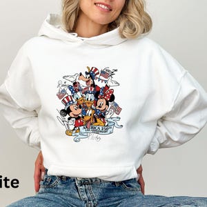 Puede incluir: Sudadera con capucha blanca con un gr&aacute;fico colorido de personajes de Disney, incluyendo Mickey y Minnie Mouse, Donald Duck y Goofy, celebrando el 250 aniversario de EE. UU. El dise&ntilde;o incorpora elementos patri&oacute;ticos como banderas y estrellas.