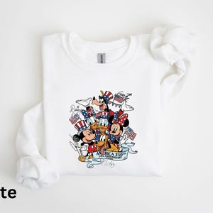 Puede incluir: Sudadera blanca de manga larga con un gr&aacute;fico colorido de personajes de Disney, incluyendo Mickey y Minnie Mouse, Donald Duck y Goofy, celebrando el 250 aniversario de EE. UU. El dise&ntilde;o incorpora elementos patri&oacute;ticos como banderas y estrellas.