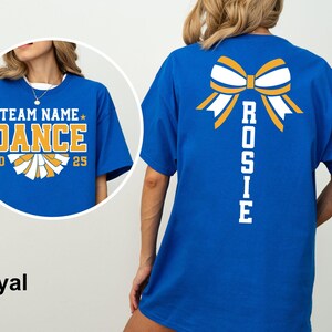 May include: Royal blue t-shirt with a cheerleading design. The front features the text "TEAM NAME DANCE 2025" in gold and white. The back displays a bow graphic and the name "ROSIE" in white.