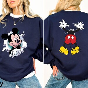 May include: Navy blue sweatshirt featuring a Mickey Mouse graphic. The front shows Mickey breaking through a white-edged hole. The back displays Mickey's full body with outstretched arms. The sweatshirt has a white crew neck.