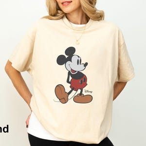 May include: Sand-colored t-shirt featuring a classic Mickey Mouse graphic. The cartoon character is depicted in his iconic pose, wearing red shorts, brown shoes, and black gloves. The Disney logo is visible at the bottom right.
