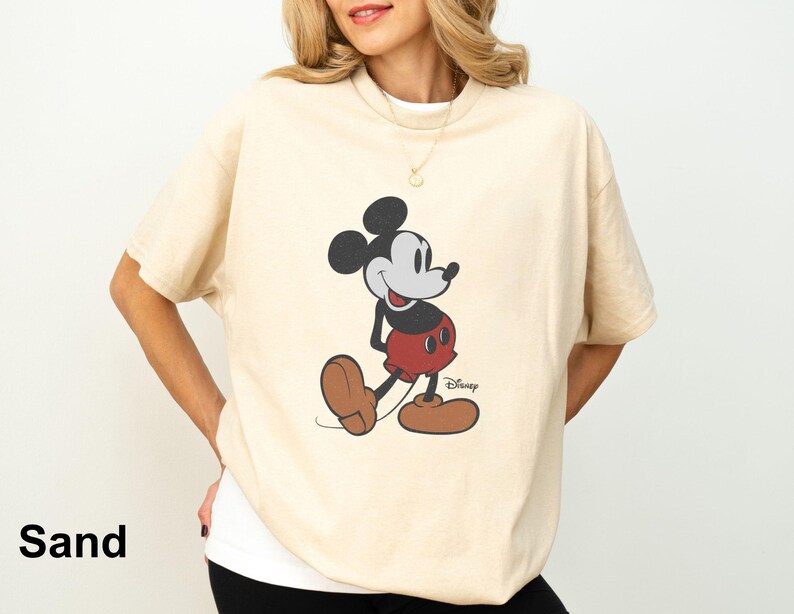 May include: Sand-colored t-shirt featuring a classic Mickey Mouse graphic. The cartoon character is depicted in his iconic pose, wearing red shorts, brown shoes, and black gloves. The Disney logo is visible at the bottom right.