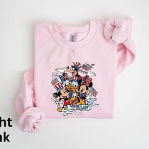 Puede incluir: Sudadera rosa claro con un gr&aacute;fico de los personajes de Disney Mickey Mouse, Minnie Mouse, Donald Duck, Daisy Duck, Goofy y Pluto con atuendos patri&oacute;ticos. El gr&aacute;fico incluye el texto "America 250th Anniversary".