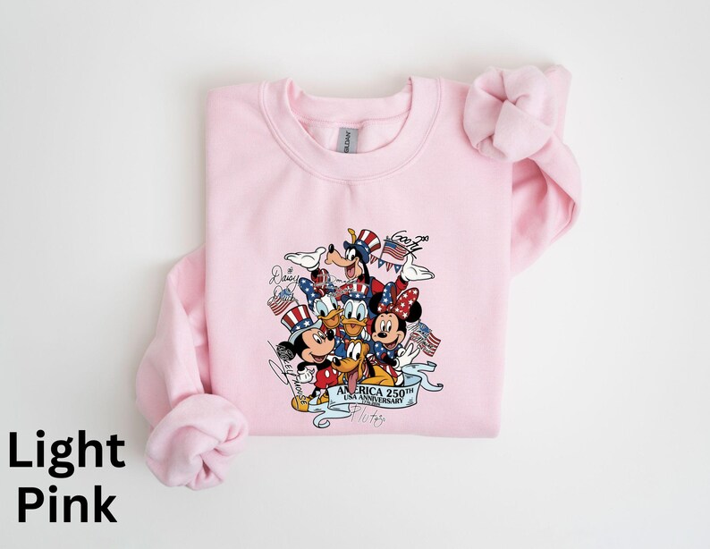 Puede incluir: Sudadera rosa claro con un gr&aacute;fico de los personajes de Disney Mickey Mouse, Minnie Mouse, Donald Duck, Daisy Duck, Goofy y Pluto con atuendos patri&oacute;ticos. El gr&aacute;fico incluye el texto "America 250th Anniversary".