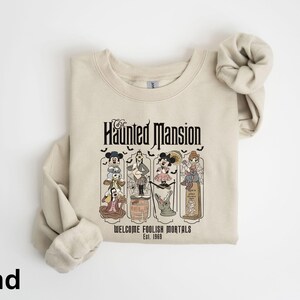May include: Sand-colored sweatshirt featuring a graphic of the Haunted Mansion with cartoon characters. The text "Haunted Mansion" is above the graphic, with "Welcome Foolish Mortals Est. 1969" below. The sweatshirt has long sleeves and a crew neck.