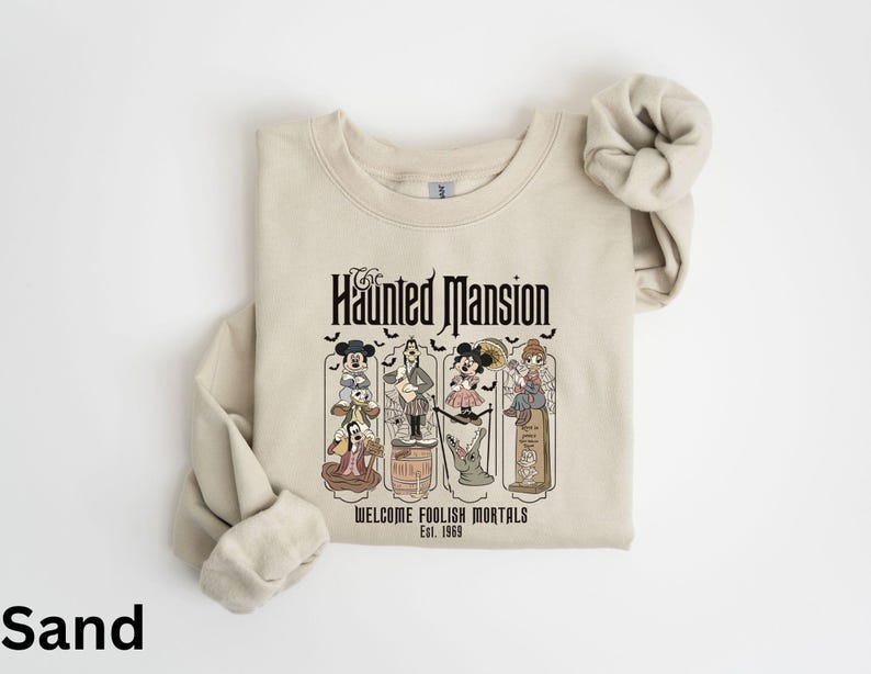 May include: Sand-colored sweatshirt featuring a graphic of the Haunted Mansion with cartoon characters. The text "Haunted Mansion" is above the graphic, with "Welcome Foolish Mortals Est. 1969" below. The sweatshirt has long sleeves and a crew neck.