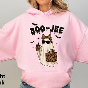 Op de afbeelding: Lichtroze hoodie met een afbeelding van een spook met een zonnebril en een luipaardstrik, met een koffiebeker en een luipaardtas. Het woord "BOO-JEE" staat boven het spook, met vleermuizen.