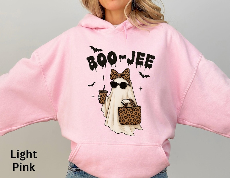 Op de afbeelding: Lichtroze hoodie met een afbeelding van een spook met een zonnebril en een luipaardstrik, met een koffiebeker en een luipaardtas. Het woord "BOO-JEE" staat boven het spook, met vleermuizen.