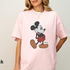 May include: A light pink t-shirt featuring a classic Mickey Mouse graphic. The cartoon character is depicted in black, red, and brown, with the Disney logo visible. The shirt has short sleeves and a relaxed fit.