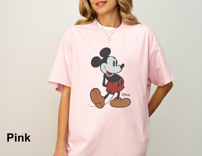May include: A light pink t-shirt featuring a classic Mickey Mouse graphic. The cartoon character is depicted in black, red, and brown, with the Disney logo visible. The shirt has short sleeves and a relaxed fit.