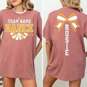 May include: Heather mauve t-shirt with "TEAM NAME DANCE 2025" in gold and white lettering on the front. The back features a bow graphic with the name "ROSIE" in white. The shirt has a crew neck and short sleeves.