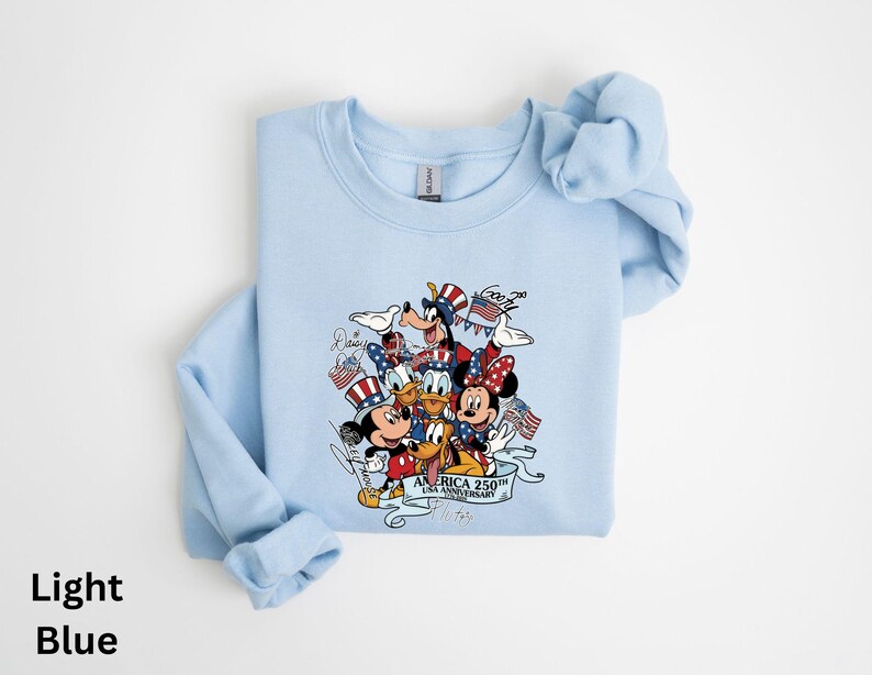 Puede incluir: Sudadera azul claro con un dise&ntilde;o de Mickey Mouse, Minnie Mouse, Donald, Goofy y Pluto con atuendos patri&oacute;ticos. El dise&ntilde;o incluye banderas americanas y el texto "America 250th Anniversary".