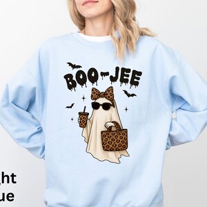 Op de afbeelding: Lichtblauwe sweatshirt met een cartoongeest met een zonnebril, een luipaardstrik en een handtas en koffiebeker met luipaardprint. De woorden "BOO-JEE" staan boven de geest. Zwarte vleermuizen en sterren maken het ontwerp compleet.